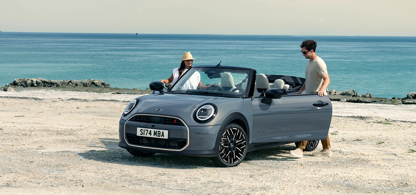 Front left view of the MINI Cooper Convertible in Copper Grey at a beach with two people next to it.