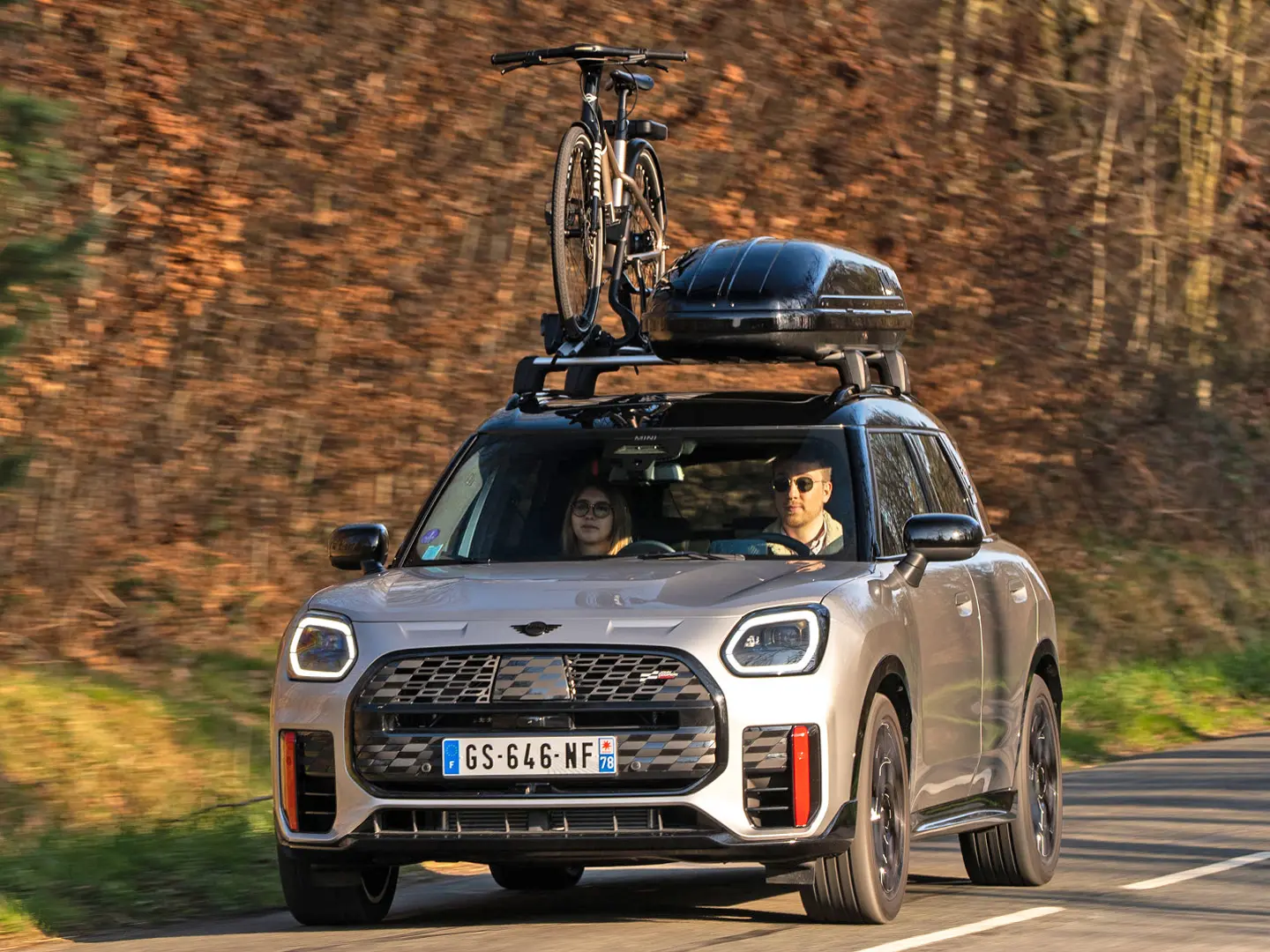 MINI Countryman Cooper in Silver on a mountain road with MINI Roof Rack, MINI Roof Box, MINI Roof Cycle Carrier with one bike mounted plus MINI John Cooper Works winglet – front view.