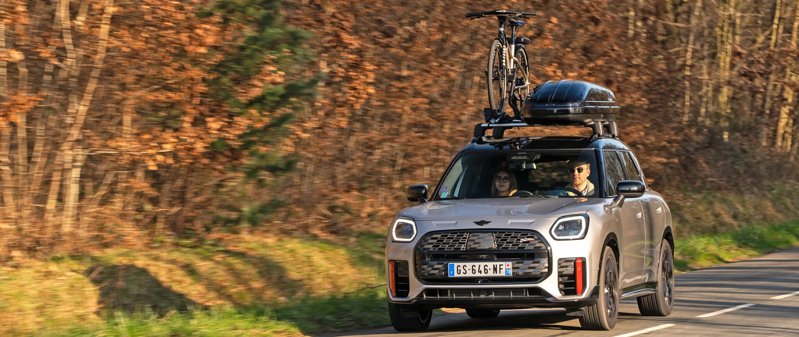 MINI Countryman Cooper in Silver on a mountain road with MINI Roof Rack, MINI Roof Box, MINI Roof Cycle Carrier with one bike mounted plus MINI John Cooper Works winglet – front view.