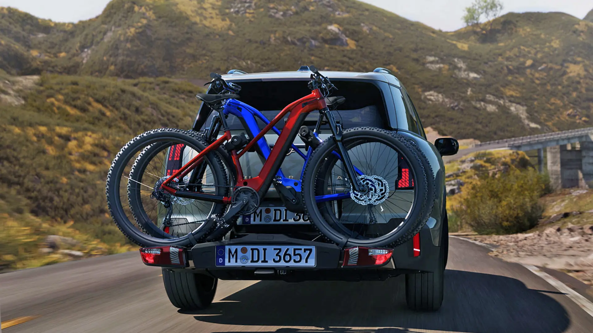 MINI Countryman in Smokey Green equipped with rear bike carrier system and two mountain bikes attached, rear view, driving along a mountain road.