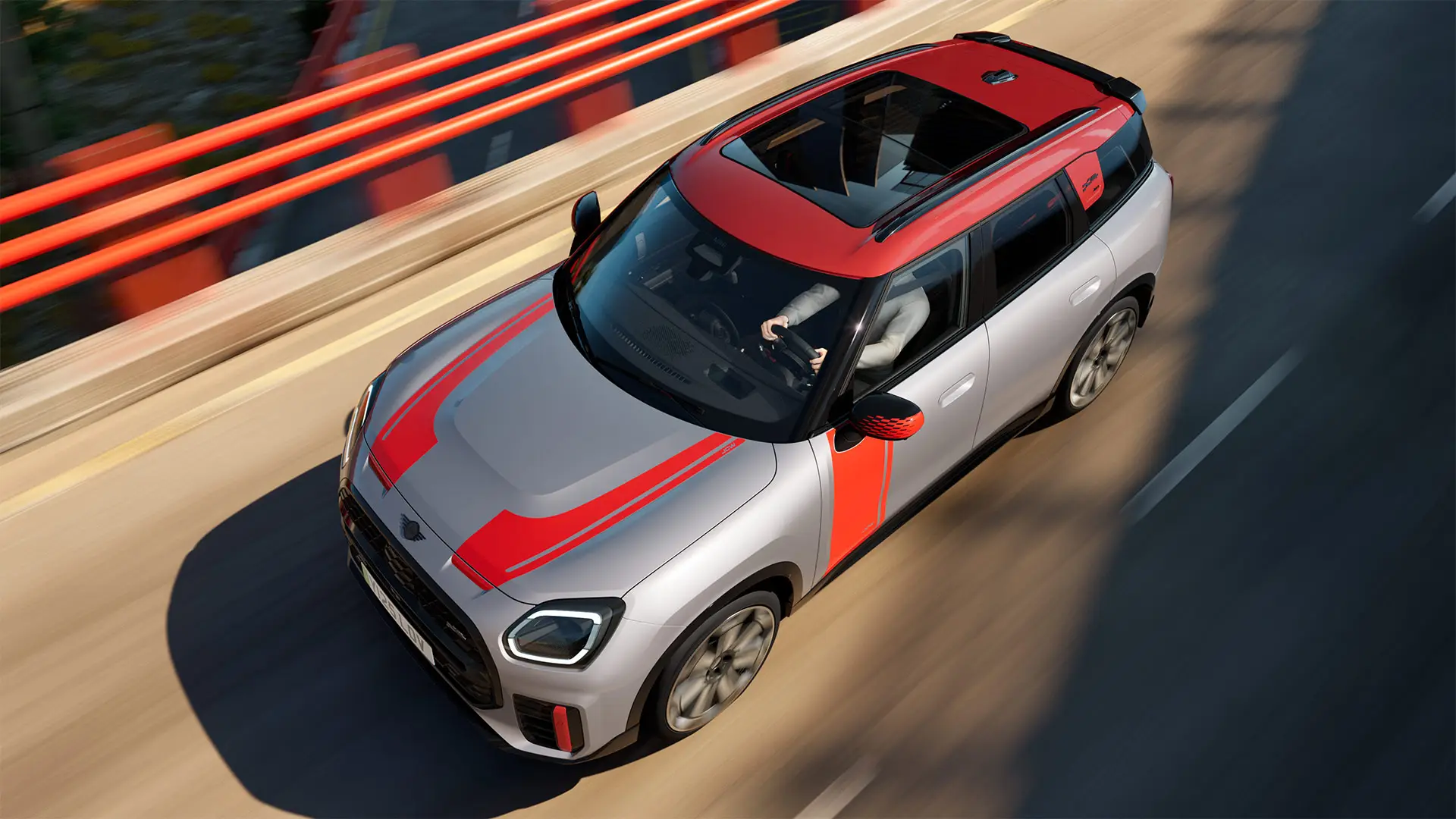 MINI Countryman John Cooper Works in Melting Silver with mirror cap, side and bonnet decals, roof, and winglet in Chili Red, driving along city street – front-side view.